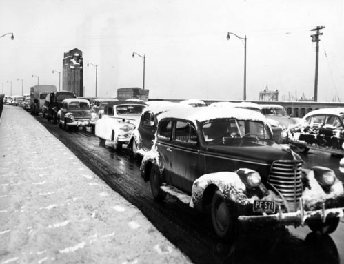 A snowy scene of traffic with vintage cars lined up on a road, featuring a clock tower in the background.