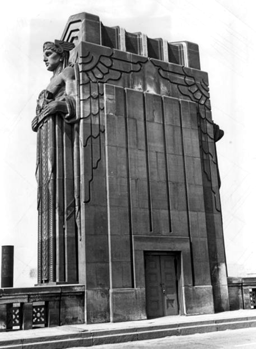 Art Deco monument featuring a large winged figure, with intricate designs and a door at the base, set against a clear sky.
