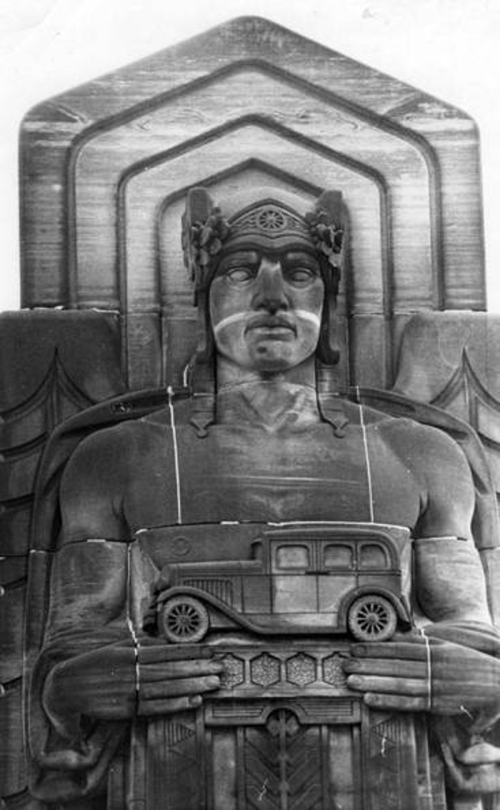 A carved statue of a muscular figure holding a vintage car, adorned with a headdress and wings, set against a decorative backdrop.