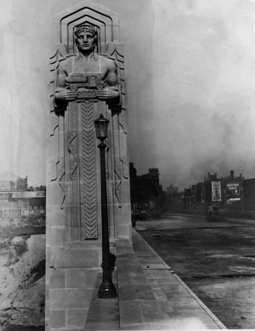 A historic black-and-white photo of a stone sculpture on a bridge, featuring a muscular figure holding a vehicle.