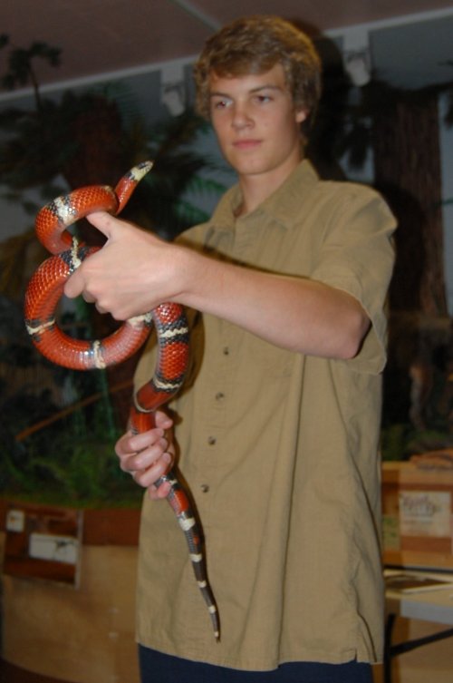 A young person holds a colorful snake, showcasing it in a natural setting with greenery in the background.