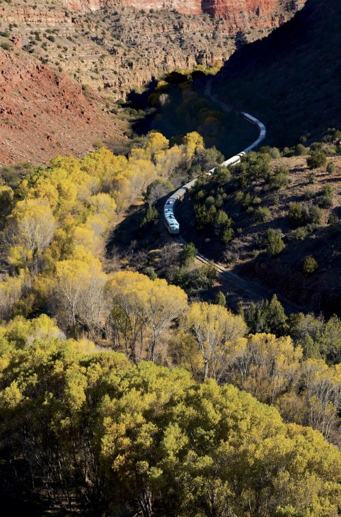 A winding train travels through a canyon surrounded by vibrant autumn foliage and rocky cliffs.
