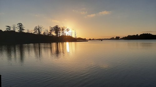 Sunset over a calm lake, with silhouettes of trees along the shore reflecting in the water.