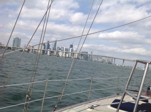 View from a sailboat on water, with a city skyline and bridge in the background under a cloudy sky.