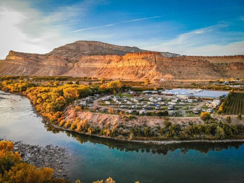 Aerial view of a campground by a river, surrounded by colorful trees and rocky cliffs under a clear blue sky.