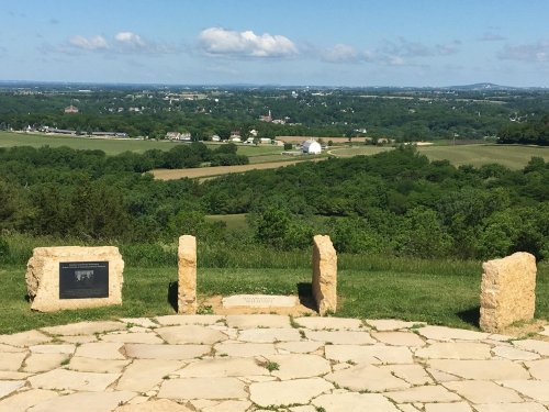 A scenic view from a hilltop, featuring stone markers and lush green fields under a blue sky with clouds.