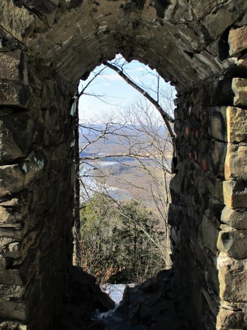 View through a stone archway, revealing a landscape of trees and a river in the distance under a clear blue sky.