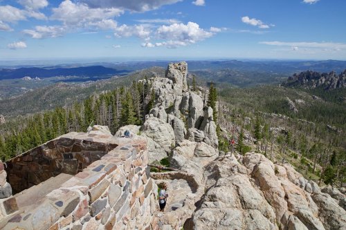 A scenic view from a rocky mountain peak, overlooking lush forests and distant hills under a blue sky with clouds.