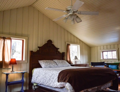 Cozy bedroom with a wooden bed, ceiling fan, and warm lighting from a lamp, featuring a rustic, inviting atmosphere.