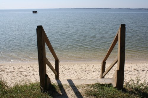 Wooden steps lead down to a calm beach with gentle waves and a distant dock on the water.