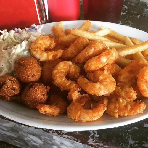 A plate of fried shrimp, hushpuppies, coleslaw, and French fries, served with drinks in the background.