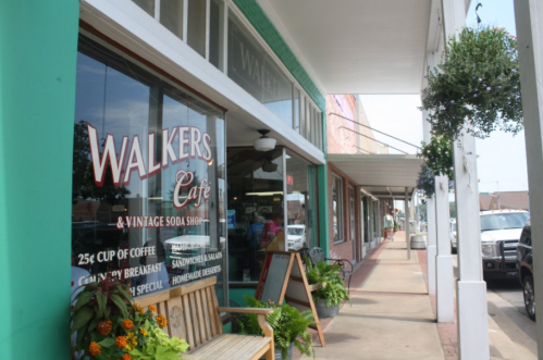 A quaint café with a vintage soda shop sign, featuring outdoor seating and hanging plants along a charming street.