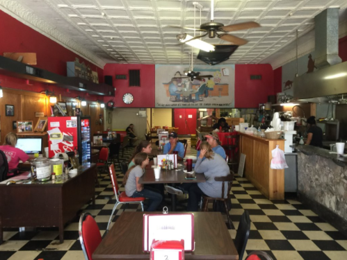 A cozy diner with red walls, checkered floors, and patrons enjoying meals at tables. A counter and kitchen are visible.