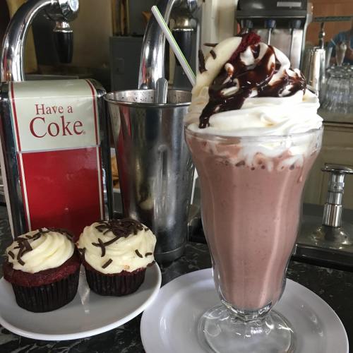 A chocolate milkshake topped with whipped cream and chocolate drizzle, alongside two red velvet cupcakes on a plate.