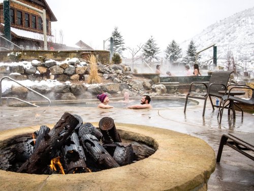 People relax in a hot spring surrounded by snow, with a fire pit in the foreground and mountains in the background.