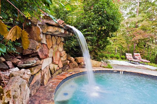 A serene backyard scene featuring a stone waterfall flowing into a circular pool, surrounded by lush greenery and lounge chairs.
