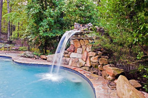 A serene pool with a natural stone waterfall surrounded by lush greenery and a stone patio.