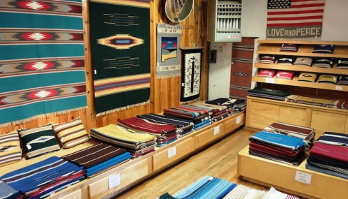 A cozy shop displaying colorful woven textiles and blankets on wooden shelves, with wall hangings and an American flag.
