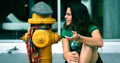 A girl sits next to a yellow fire hydrant wearing a hat and scarf, engaged in a conversation.