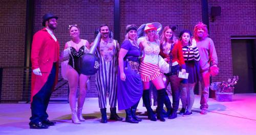 A group of people in colorful costumes posing together on stage under purple lighting.