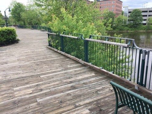 A wooden walkway along a river, lined with green trees and a railing, with benches visible in the foreground.