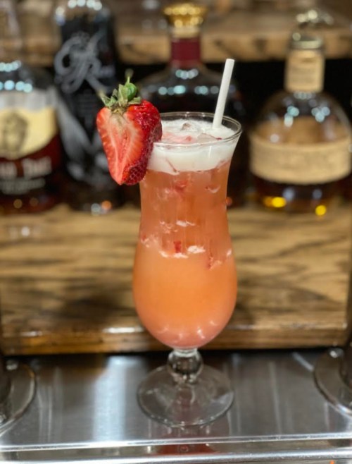 A refreshing cocktail in a tall glass, garnished with a strawberry, set against a backdrop of liquor bottles.