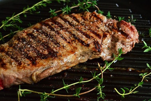 Grilled steak with grill marks, garnished with fresh thyme and sprinkled with black pepper on a dark background.
