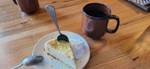 A slice of key lime pie with whipped cream on a plate, next to a brown mug of coffee on a wooden table.
