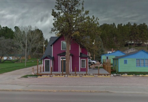 A colorful two-story house with a dark roof, surrounded by trees and other small buildings, under a cloudy sky.