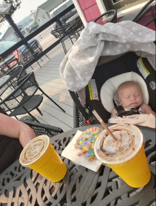 A sleeping baby in a stroller beside two yellow cups and a cookie on a table at an outdoor café.
