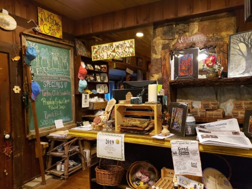 Cozy café interior with a chalkboard menu, wooden decor, and various handmade items displayed on shelves.