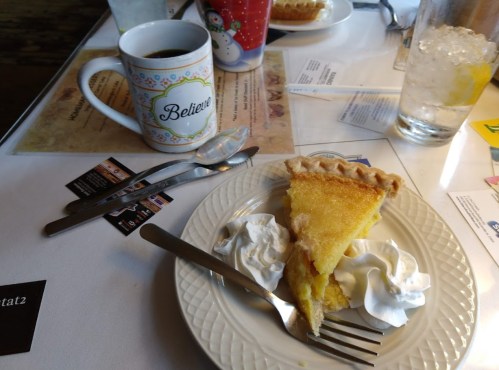 A slice of pie with whipped cream on a plate, a coffee mug, and a glass of water on a table.