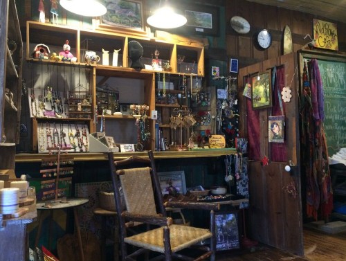 A cozy shop interior filled with handmade jewelry, crafts, and rustic decor, featuring a wooden chair and shelves.