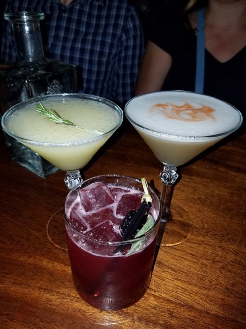 Three cocktails on a wooden table: two martinis with garnishes and a dark red drink with ice and a straw.