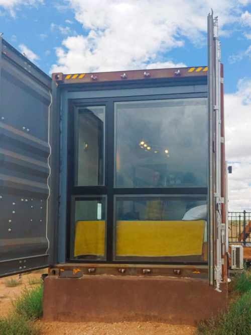 A modern shipping container converted into a cozy living space with large windows and a yellow bedspread.