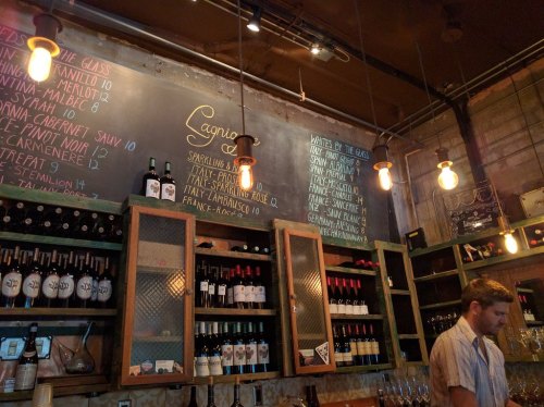 A cozy bar with a chalkboard menu, shelves of wine bottles, and warm pendant lights. A bartender is working.