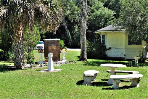 A small memorial with flowers and benches, surrounded by trees and a quaint yellow building in a grassy area.
