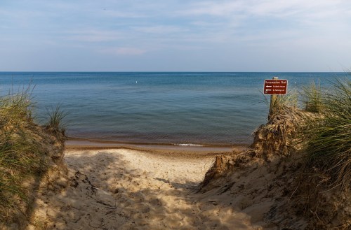 A sandy path leads to a calm lake, with grass on either side and a warning sign in the foreground.