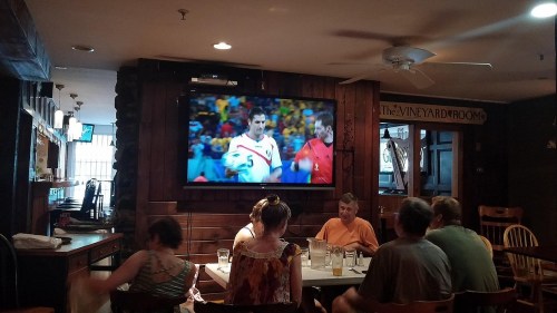 A sports bar scene with patrons watching a soccer game on a large TV screen. Tables are filled with drinks and food.