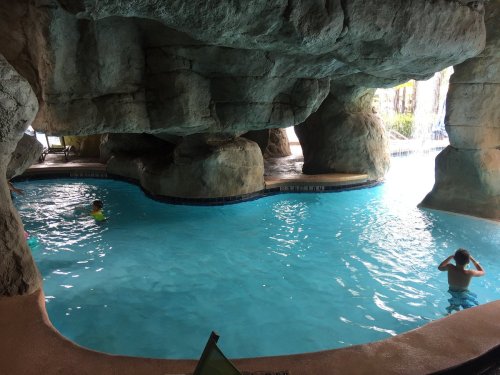 Two children swim in a blue pool surrounded by rocky formations and a waterfall in a cave-like setting.