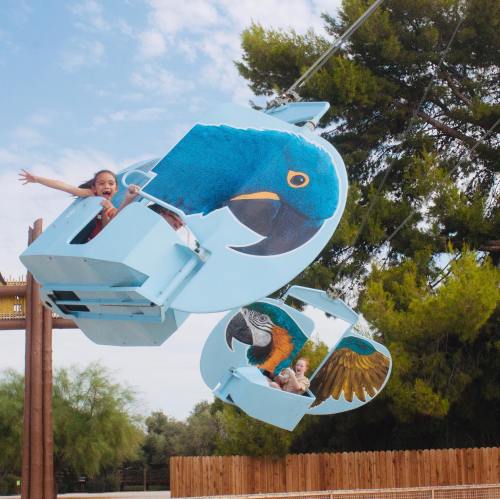 Two children enjoy a colorful bird-themed ride, smiling and waving as they soar above a park.