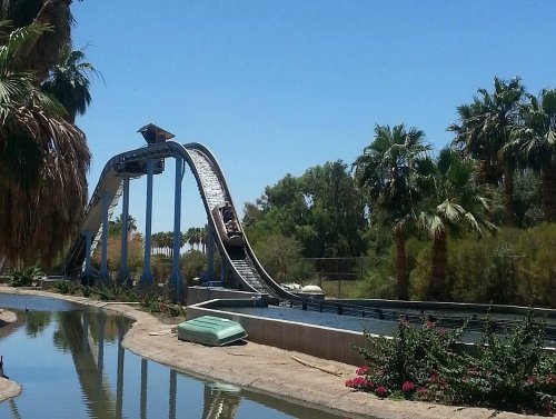 A water ride with a steep drop, surrounded by palm trees and a calm waterway under a clear blue sky.