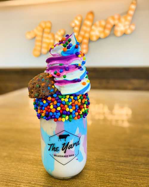 A colorful milkshake topped with swirled whipped cream, a cookie, and rainbow sprinkles in a branded glass.