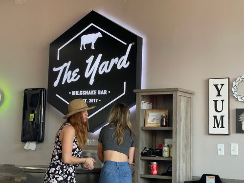 Two women converse in front of a large illuminated sign for "The Yard Milkshake Bar" in a cozy interior setting.