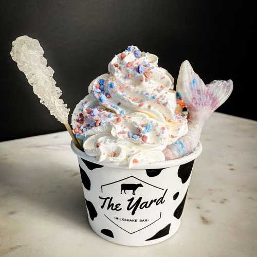 A cup of colorful whipped cream topped with sprinkles and a mermaid tail, served in a cow-print container.