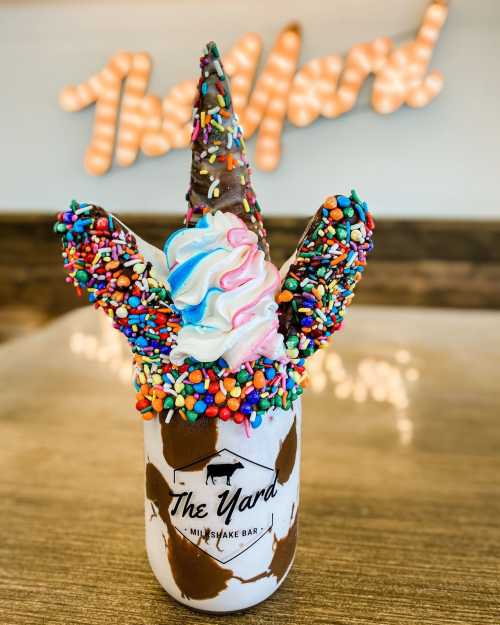 A colorful ice cream treat in a jar, topped with whipped cream, chocolate, and rainbow sprinkles, with a cone and candy accents.