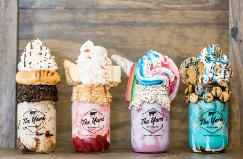 Four colorful milkshakes topped with whipped cream, cookies, and sweets, served in mason jars with "The Yard" logo.