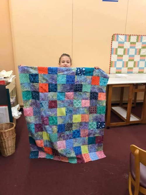 A child holds up a colorful patchwork quilt in a room with crafting supplies and a table in the background.