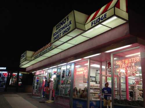 A brightly lit sweet shop at night, featuring signs for fudge, taffy, and homemade candies.