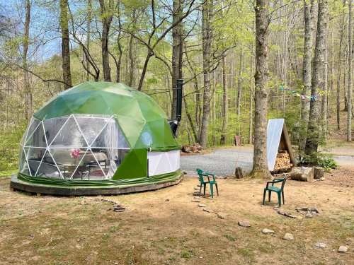 A green geodesic dome cabin surrounded by trees, with two chairs and a firewood stack nearby.